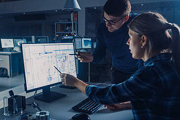Two people sit in front of a computer and work on a CAD model.